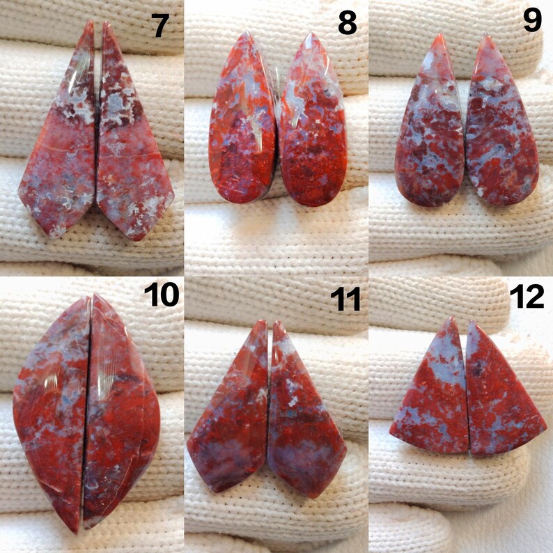 May include: A collection of six pairs of polished gemstone cabochons. Each pair features a unique shape and pattern, with a mix of red, gray, and white hues. The gemstones are displayed on a white, textured surface.