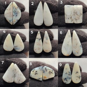 May include: A collection of nine pairs of polished dendritic opal cabochons. The stones vary in shape, including teardrop, rectangular, and irregular forms. Each stone displays a creamy white base color with dendritic inclusions in shades of black, gray, and blue.