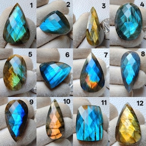 Puede incluir: Una colección de doce piedras preciosas de labradorita facetadas, cada una con formas únicas y patrones de color iridiscentes. Las piedras muestran una gama de azules, verdes y amarillos, con algunas piedras que muestran un destello naranja. Las piedras se muestran sobre una superficie blanca.