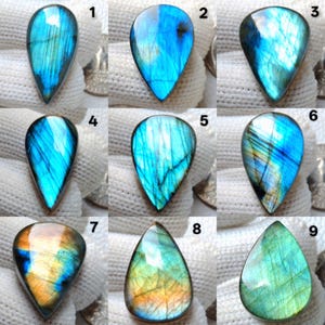 May include: Nine teardrop-shaped labradorite gemstones, each displaying iridescent flashes of blue, green, and gold. The stones, with unique patterns, are presented on a white surface. The gemstones' colours are highlighted by the white background.