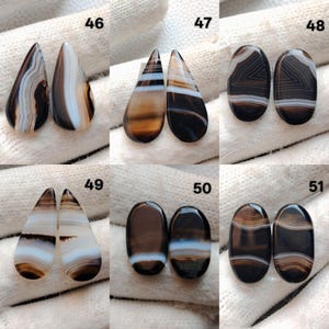 May include: A selection of polished agate gemstones in diverse shapes, such as teardrop and oval. The stones showcase a spectrum of colours, including brown, black, and white, with banded patterns. The gemstones are presented on a white surface.