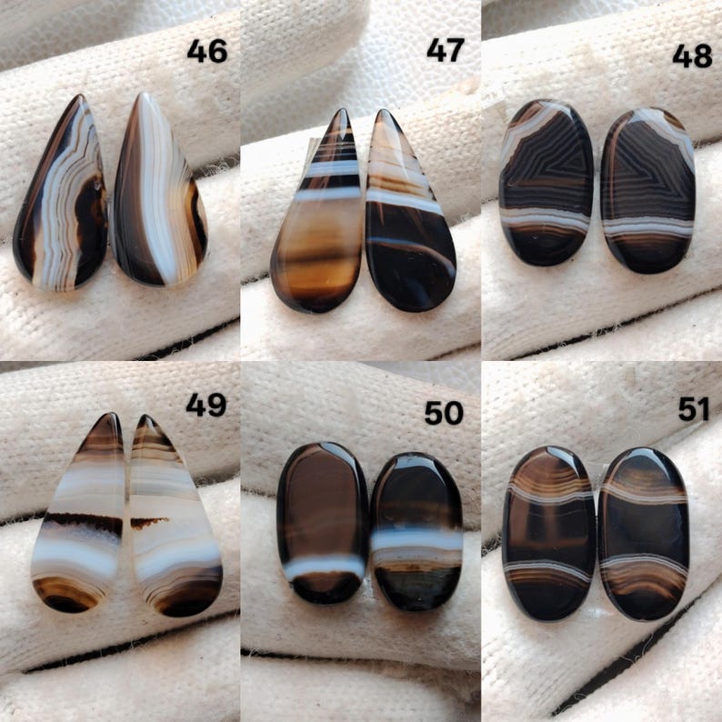 May include: A selection of polished agate gemstones in diverse shapes, such as teardrop and oval. The stones showcase a spectrum of colours, including brown, black, and white, with banded patterns. The gemstones are presented on a white surface.