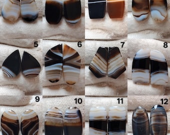 May include: A collection of twelve pairs of polished agate gemstones, each with unique patterns and colors. The stones vary in shape, including teardrop and oval, and feature bands of brown, white, and black. The gemstones are displayed on a white surface.