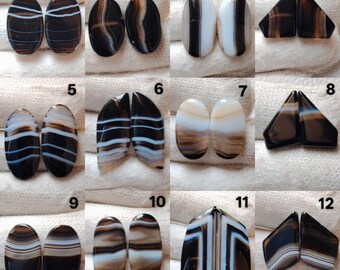 May include: A collection of twelve pairs of polished agate gemstones in various shapes and patterns. The stones feature bands of brown, black, white, and tan colors. Some are oval, others triangular or irregular.