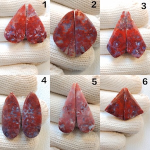 May include: A collection of six pairs of polished gemstones, each split into two pieces. The stones display a mix of red, blue, and white hues. The shapes vary, including teardrop, diamond, and triangular forms, suitable for jewelry making.