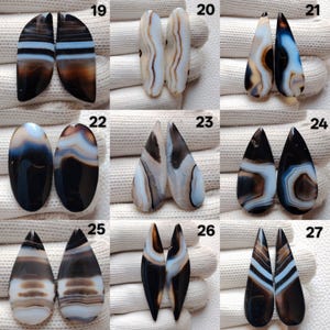 May include: A selection of polished agate gemstone pairs in diverse shapes, including oval, teardrop, and elongated forms. The stones exhibit a spectrum of colours, such as brown, black, white, and blue, with banded patterns. The gemstones are displayed on a white surface.