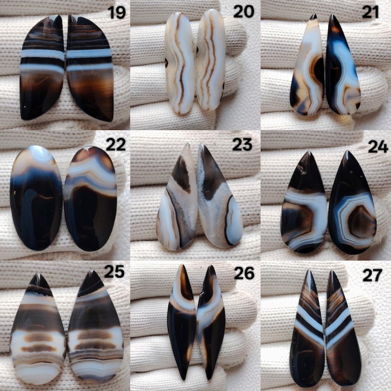 May include: A selection of polished agate gemstone pairs in diverse shapes, including oval, teardrop, and elongated forms. The stones exhibit a spectrum of colours, such as brown, black, white, and blue, with banded patterns. The gemstones are displayed on a white surface.