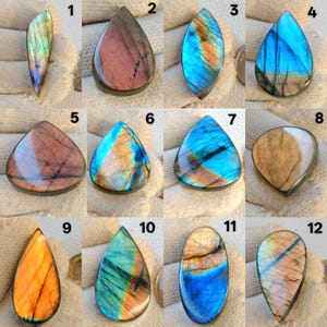 Puede incluir: Una colección de doce piedras preciosas de labradorita pulidas. Las piedras presentan diversas formas, incluyendo lágrimas, óvalos y triángulos. Los colores varían, con azules, naranjas, verdes y morados, y destellos iridiscentes. Cada una tiene patrones únicos.