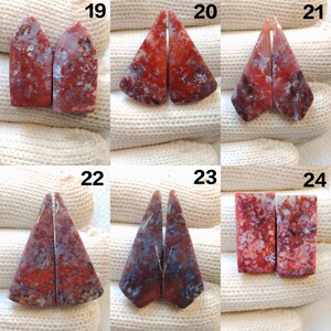 May include: A collection of six pairs of polished gemstones, each with a unique shape and pattern. The stones display a mix of red, white, and blue hues, with visible veining and marbling. The gemstones are arranged on a white, textured surface.