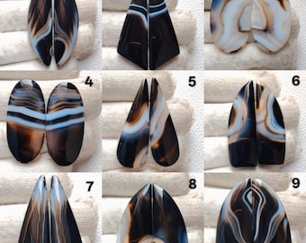May include: Nine pairs of polished agate gemstones in various shapes, including oval, triangular, and teardrop. The stones feature a mix of black, brown, white, and blue banding patterns. Each pair is split down the middle, showcasing the symmetrical designs.