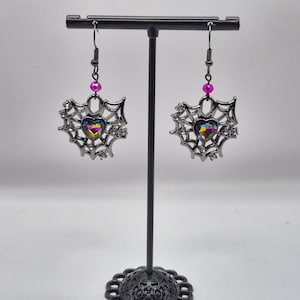 May include: A pair of dangle earrings featuring a dark metallic spiderweb frame with a central iridescent heart-shaped stone. Each earring is accented with a small bright pink bead above the web and clear crystals embedded along the outer edges.