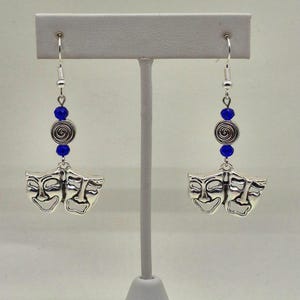 Op de afbeelding: Zilverkleurige oorhangers met komedie- en tragediemaskers. Elke oorbel heeft een blauwe kraal, een zilveren spiraal en een paar maskers. De maskers zijn gedetailleerd met gezichtsuitdrukkingen. De oorbellen hangen aan zilveren haken.