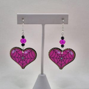 May include: A pair of heart-shaped earrings featuring a pink spiderweb design. The earrings have a black and gold trim, with a small blue heart at the web's centre. They hang from silver hooks with black and purple beads.