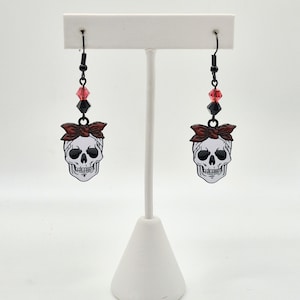 Könnte beinhalten: Ein Paar schwarze Schädelohrringe mit roten und schwarzen Akzenten. Jeder Ohrring zeigt einen weißen Schädel mit roter Schleife, der an einem schwarzen Haken mit roten und schwarzen Perlen hängt. Die Ohrringe werden auf einem weißen Ständer präsentiert.