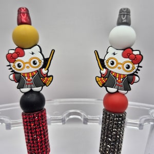 Peut inclure: Deux stylos avec un design Hello Kitty déguisé en sorcier. Chaque stylo a une combinaison de couleurs différente, avec une base rouge et noire, et une base jaune et blanche. Les stylos sont décorés de strass.