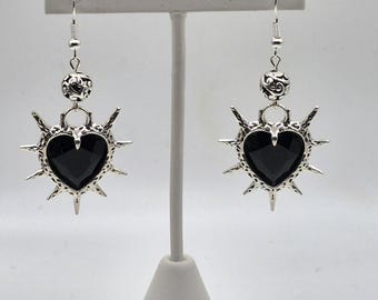 May include: A pair of silver-toned earrings. Each earring features a black heart-shaped stone surrounded by a spiky, sunburst design. Above the heart is a decorative bead and a hook for the ear.