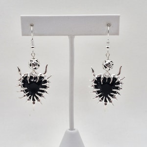 Może przedstawiać: Para srebrnych kolczyków z czarnym sercem w środku. Kolczyki mają kolczasty, ozdobny wzór wokół serca i dekoracyjną kulkę na górze. Wyeksponowane na białym stojaku.