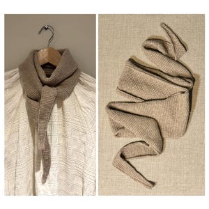 以下が含まれることがあります： ユニークなデザインのベージュ色のニットスカーフ。ハンガーに掛けられた状態と、広げられた状態の両方で表示されています。スカーフは質感があり、長く尖った端があります。ニュートラルな色合いで、汎用性の高いアクセサリーです。