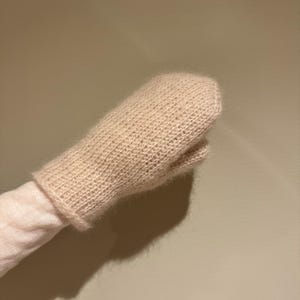 May include: A light beige knitted mitten with a matching cuff. The mitten is made of a soft, textured yarn and has a classic design. The thumb is separate from the rest of the mitten. The cuff is a lighter shade of beige.