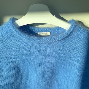 Può includere: Un maglione blu chiaro girocollo realizzato in un materiale morbido e soffice. Il maglione è appeso a una gruccia bianca. L'etichetta recita "moffeloff".