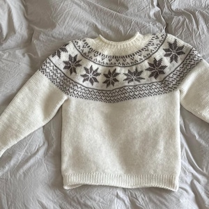 May include: A white knit sweater with a crew neck and long sleeves. The sweater features a gray snowflake pattern across the chest and upper sleeves. The snowflake design is repeated around the neckline. The sweater is lying on a wrinkled white sheet.
