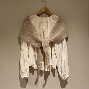 Könnte beinhalten: Cremefarbene Bluse mit langen Ärmeln und strukturiertem Muster, kombiniert mit einem beigefarbenen Strickschal, der vorne gebunden ist. Die Bluse hängt an einem Holzbügel vor einer neutralen Wand. Der Gesamteindruck ist weich und neutral.