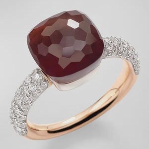May include: A gemstone ring featuring a large, faceted, dark red stone. The ring has a rose gold band with a section of small, sparkling white stones on each side of the central stone. The ring is set against a light background.