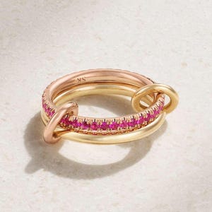 Pode incluir: Três anéis de ouro interligados, um com uma faixa de pedras preciosas rosa. Os anéis são em tons de ouro e ouro rosa. As pedras preciosas são de uma cor rosa vibrante. Os anéis são exibidos em uma superfície clara.