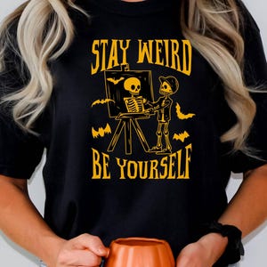 以下が含まれることがあります： 「STAY WEIRD BE YOURSELF」の黄色い文字が入った黒いTシャツ。Tシャツには、キャンバスに骸骨を描く骸骨のアーティストが描かれており、背景にはコウモリがいます。その人はオレンジ色のカボチャマグを持っています。
