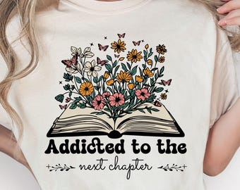 Puede incluir: Camiseta beige con un diseño de libro abierto, adornada con flores y mariposas de colores. El texto "Addicted to the next chapter" está impreso en negro. Un diseño floral está en la parte inferior.