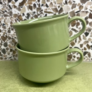May include: Two stacked green ceramic mugs with handles. The mugs are a muted shade of green and have a rounded shape.
