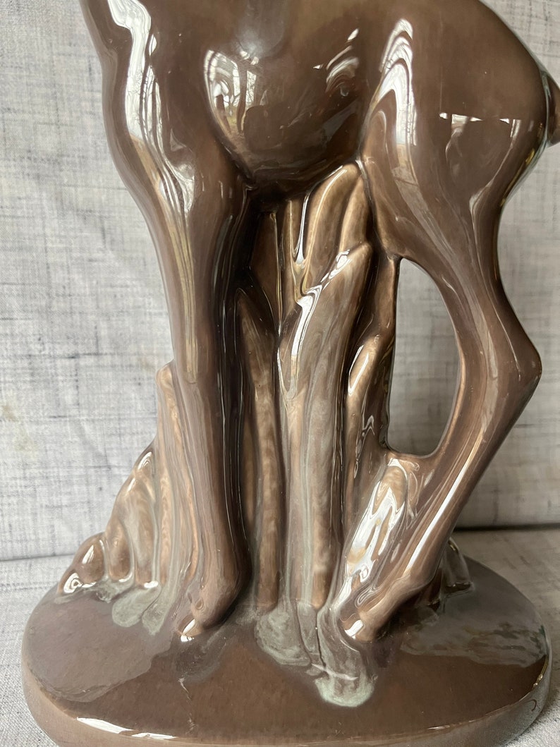 May include: A brown ceramic deer figurine standing on a base with a leafy design. The deer is glazed with a glossy finish.
