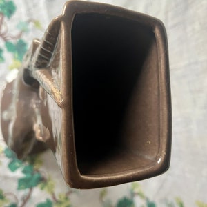 May include: A brown ceramic vase with a unique, abstract design. The vase has a wide opening and a narrow base.