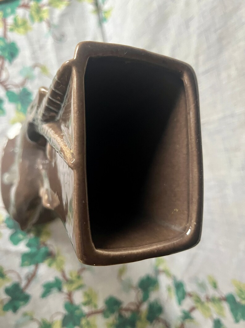 May include: A brown ceramic vase with a unique, abstract design. The vase has a wide opening and a narrow base.