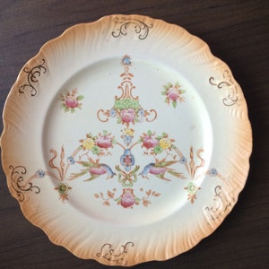 May include: A white ceramic plate with a scalloped edge and a gold floral design. The plate has a light brown glaze and features a central floral motif with birds.