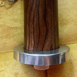 May include: A wooden dowel with a silver metal ring at the bottom. The dowel has a dark brown wood grain pattern.