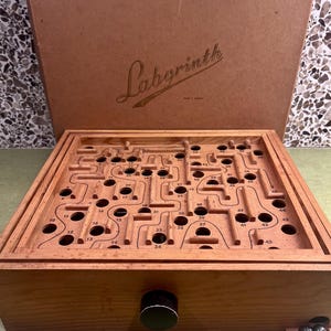 May include: A wooden Labyrinth game board featuring a maze with numbered holes and a black control knob on the front. The game includes a matching cardboard box lid with the word Labyrinth printed in script. Two metal balls sit in a small tray.