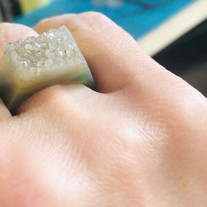 K&ouml;nnte beinhalten: Ein quadratischer Ring mit einer klaren, hellgr&uuml;nen Harzbasis und einer oberen Schicht aus wei&szlig;en, glitzernden Kristallen.