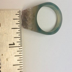 K&ouml;nnte beinhalten: Ein transparenter Harzring mit einem hellblauen und wei&szlig;en Agatstein, der an der Seite eingebettet ist. Der Ring hat einen Durchmesser von etwa 2,5 cm.