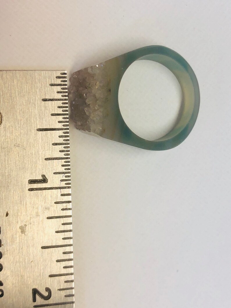 K&ouml;nnte beinhalten: Ein transparenter Harzring mit einem hellblauen und wei&szlig;en Agatstein, der an der Seite eingebettet ist. Der Ring hat einen Durchmesser von etwa 2,5 cm.