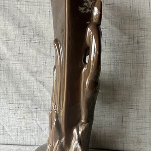 May include: A brown ceramic vase with a unique, abstract design. The vase has a tall, rectangular body with a wide base and a narrow opening. The design features a series of raised, organic shapes that resemble flames or leaves.