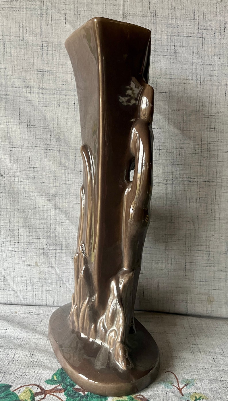 May include: A brown ceramic vase with a unique, abstract design. The vase has a tall, rectangular body with a wide base and a narrow opening. The design features a series of raised, organic shapes that resemble flames or leaves.