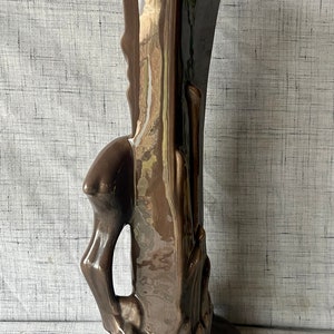 May include: A brown ceramic vase with a unique, abstract design. The vase has a tall, slender shape with a wide base and a narrow opening. The design features a stylized tree trunk with branches and leaves.