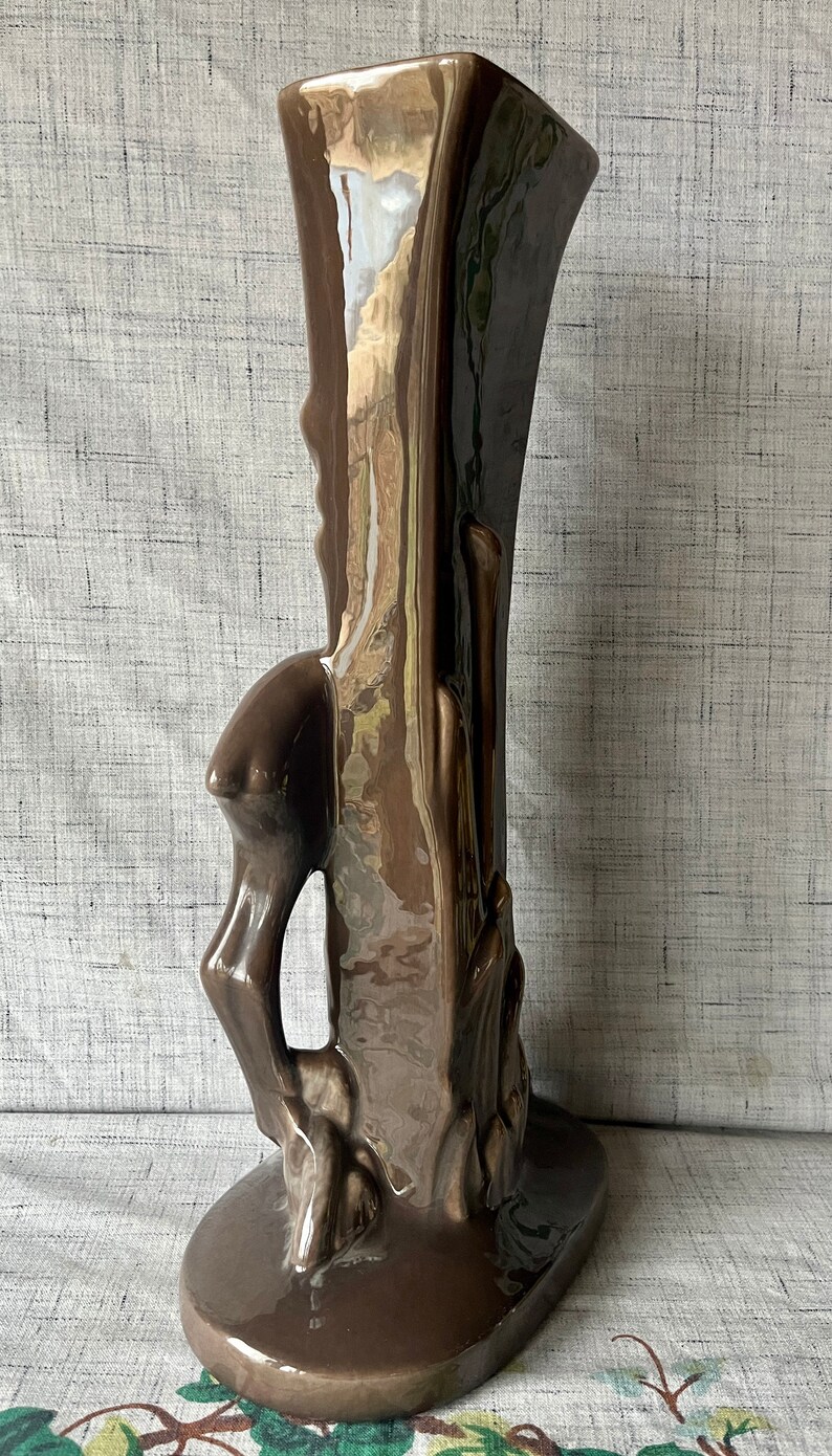 May include: A brown ceramic vase with a unique, abstract design. The vase has a tall, slender shape with a wide base and a narrow opening. The design features a stylized tree trunk with branches and leaves.