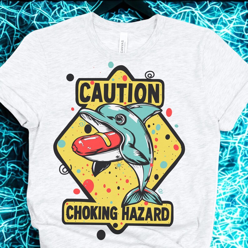 May include: White t-shirt featuring a cartoon dolphin with a hot dog in its mouth, set against a yellow diamond with the words "CAUTION" and "CHOKING HAZARD". The dolphin is teal, and the hot dog is red.