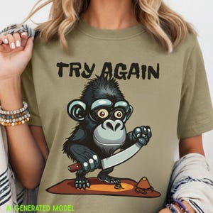 Pode incluir: Uma t-shirt verde-oliva com uma ilustração gráfica de um macaco preto segurando uma faca grande. Acima do macaco, o texto "TRY AGAIN" está impresso em letras pretas em negrito. O macaco está sobre uma forma oval marrom com um pequeno cone laranja.