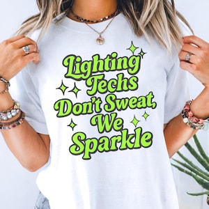 May include: A white t-shirt featuring the text "Lighting Techs Don't Sweat, We Sparkle" in a bold, lime green font with a dark outline. The design is accented with small, four-pointed star shapes in the same lime green colour, centred on the chest.