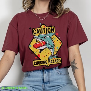 May include: A maroon t-shirt featuring a graphic of a teal dolphin with a red hot dog in its mouth. The design is set against a yellow diamond background with black text that reads CAUTION CHOKING HAZARD, surrounded by small colorful dots.