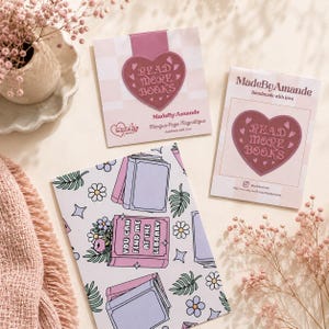 Peut inclure: Trois marque-pages décoratifs sur le thème des livres. Deux marque-pages présentent un motif en forme de cœur avec le texte "READ MORE BOOKS". Le troisième marque-page a une illustration de livre avec le texte "YOU CAN FIND ME AT THE LIBRARY".
