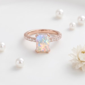Könnte beinhalten: Roségoldener Ring mit einem rechteckigen Opal-Edelstein. Das Band ist mit kleinen, klaren Steinen besetzt. Der Opal zeigt ein Farbspiel, darunter Blau, Grün und Gelb. Der Ring liegt auf weißem Grund mit Perlen und einer weißen Blume.